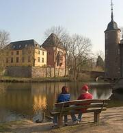 Das Foto zeigt eine Ansicht auf Schloß Burgau. Ein junges Paar sitzt am Weiher.